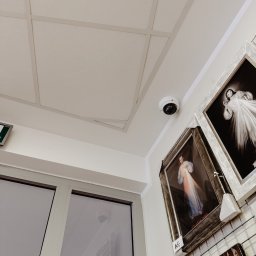 P.H.U. CamVis - systemy zabezpieczeń - Biała kamera kopułkowa zamontowana na suficie w pomieszczeniu, widoczna z perspektywy żabiej, obok wiszą obrazy w ramach na metalowej siatce.