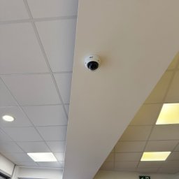 P.H.U. CamVis - systemy zabezpieczeń - Kamera kopułkowa BCS zamontowana na suficie podwieszanym w jasnym pomieszczeniu biurowym, widok z dołu, obok widoczny znak ewakuacyjny.