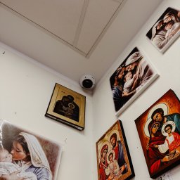 P.H.U. CamVis - systemy zabezpieczeń - Kamera kopułkowa dyskretnie zamontowana w rogu pomieszczenia, otoczona religijnymi obrazami na ścianie. Niewielki, biały korpus kamery wtapia się w tło.