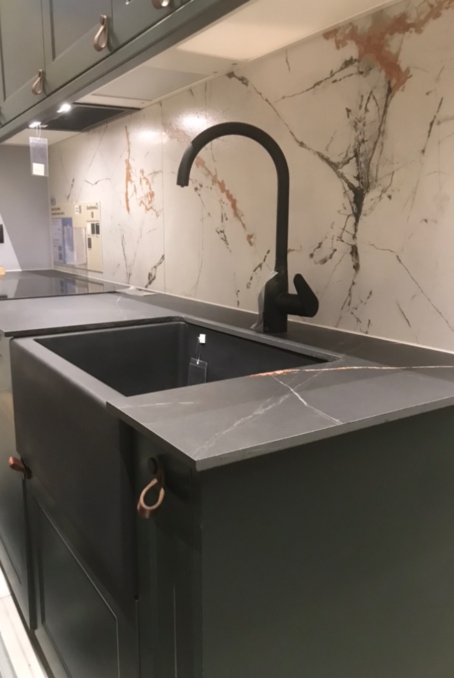 Stylowa, ciemnozielona szafka zlewozmywakowa z czarną baterią na tle marmurowej ściany. Miedziane uchwyty dodają elegancji.