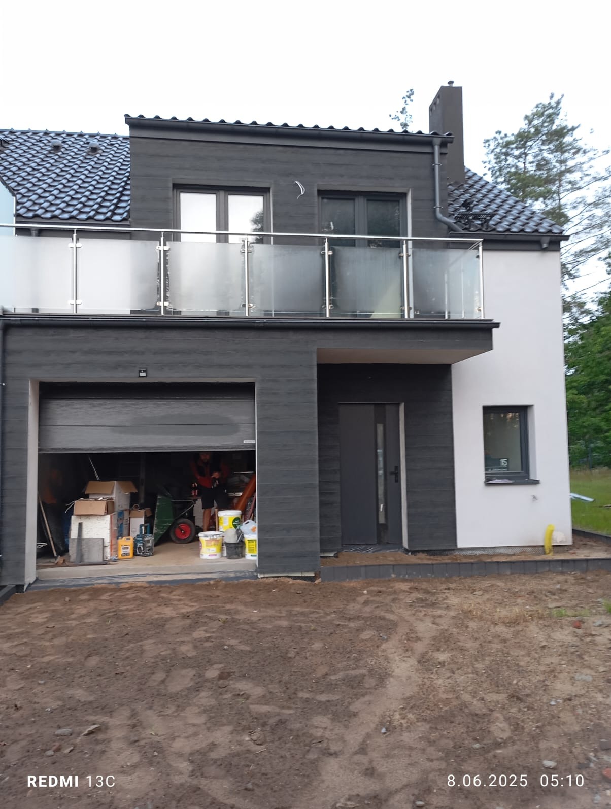 Dwukondygnacyjny dom z balkonem i garażem w trakcie wykończenia. Widoczne materiały budowlane i narzędzia w otwartym garażu. Elewacja w odcieniach szarości i bieli.