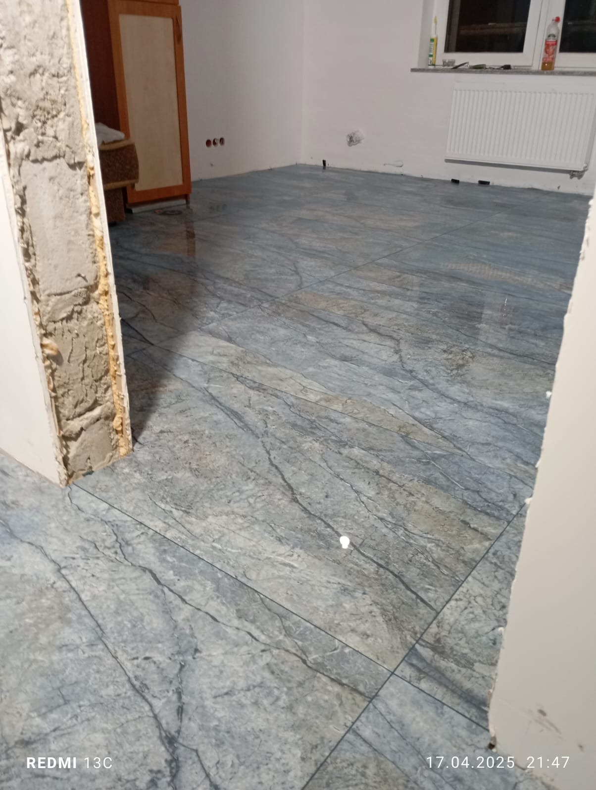 Ułożona podłoga z płytek imitujących marmur w jasnym pomieszczeniu. Widoczne ślady po zaprawie przy futrynie. Grzejnik pod oknem.