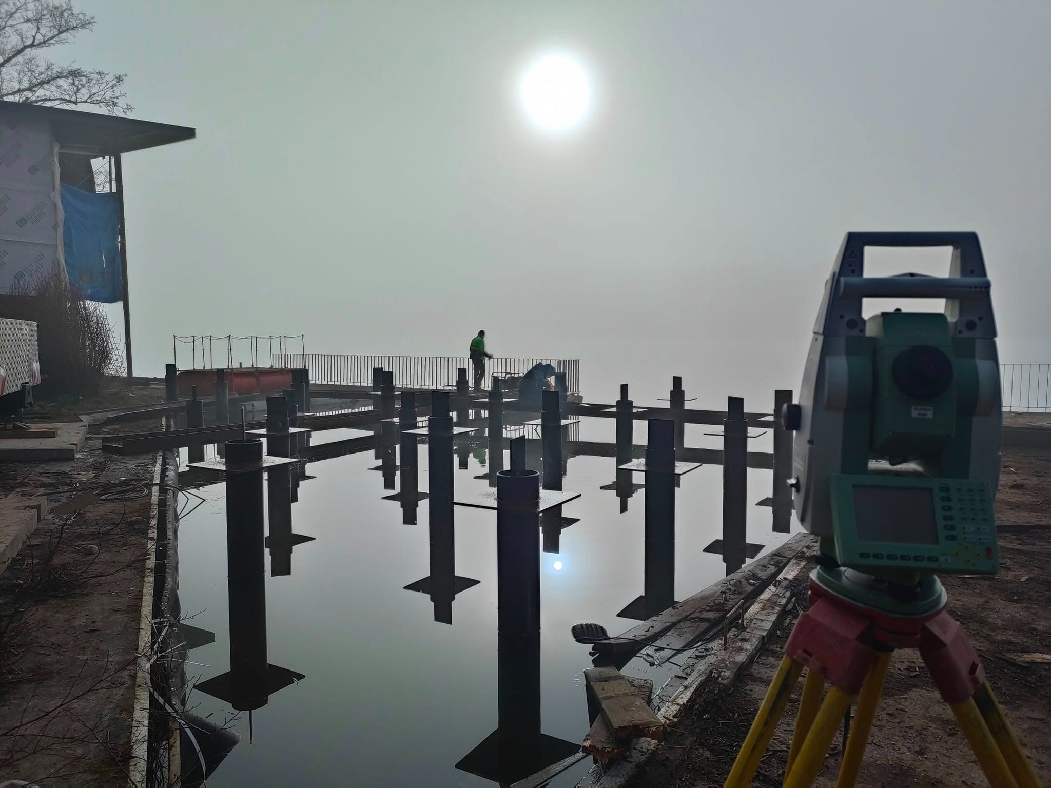 Prace pomiarowe na budowie w mglisty dzień. Teodolit na statywie, w tle metalowe słupy w wodzie i pracownik na konstrukcji. Refleksy w wodzie.