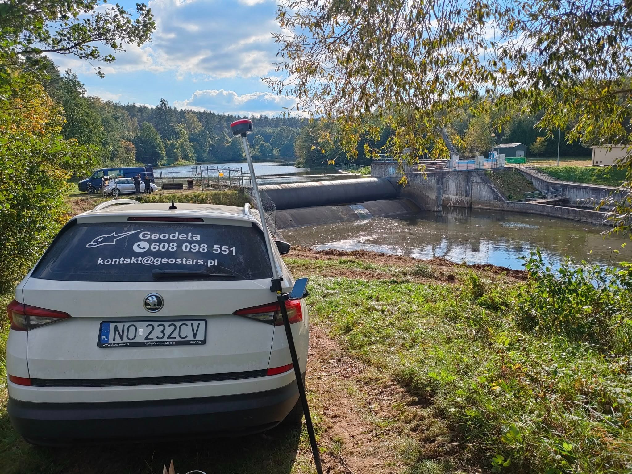 Pomiar geodezyjny przy zaporze wodnej: biała Skoda z naklejką firmy, tyczka geodezyjna, w tle woda i budowla hydrotechniczna w słoneczny dzień.