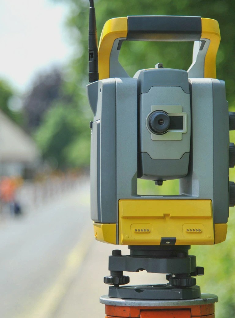 Profesjonalny teodolit geodezyjny w kolorze szaro-żółtym, ustawiony na statywie na tle rozmytej drogi i zieleni. Sprzęt gotowy do pomiarów.