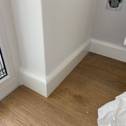 Volodymyr Shelest - Zamknięty kadr na fragment wnętrza: brązowe panele podłogowe, białe listwy przypodłogowe przy ścianie i oknie. Widoczny fragment gniazdka elektrycznego.