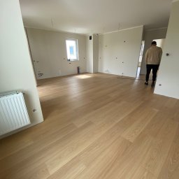 Volodymyr Shelest - Jasne panele podłogowe w nowym, przestronnym pokoju z grzejnikiem i widocznymi instalacjami elektrycznymi. Osoba w tle przechodzi przez drzwi.