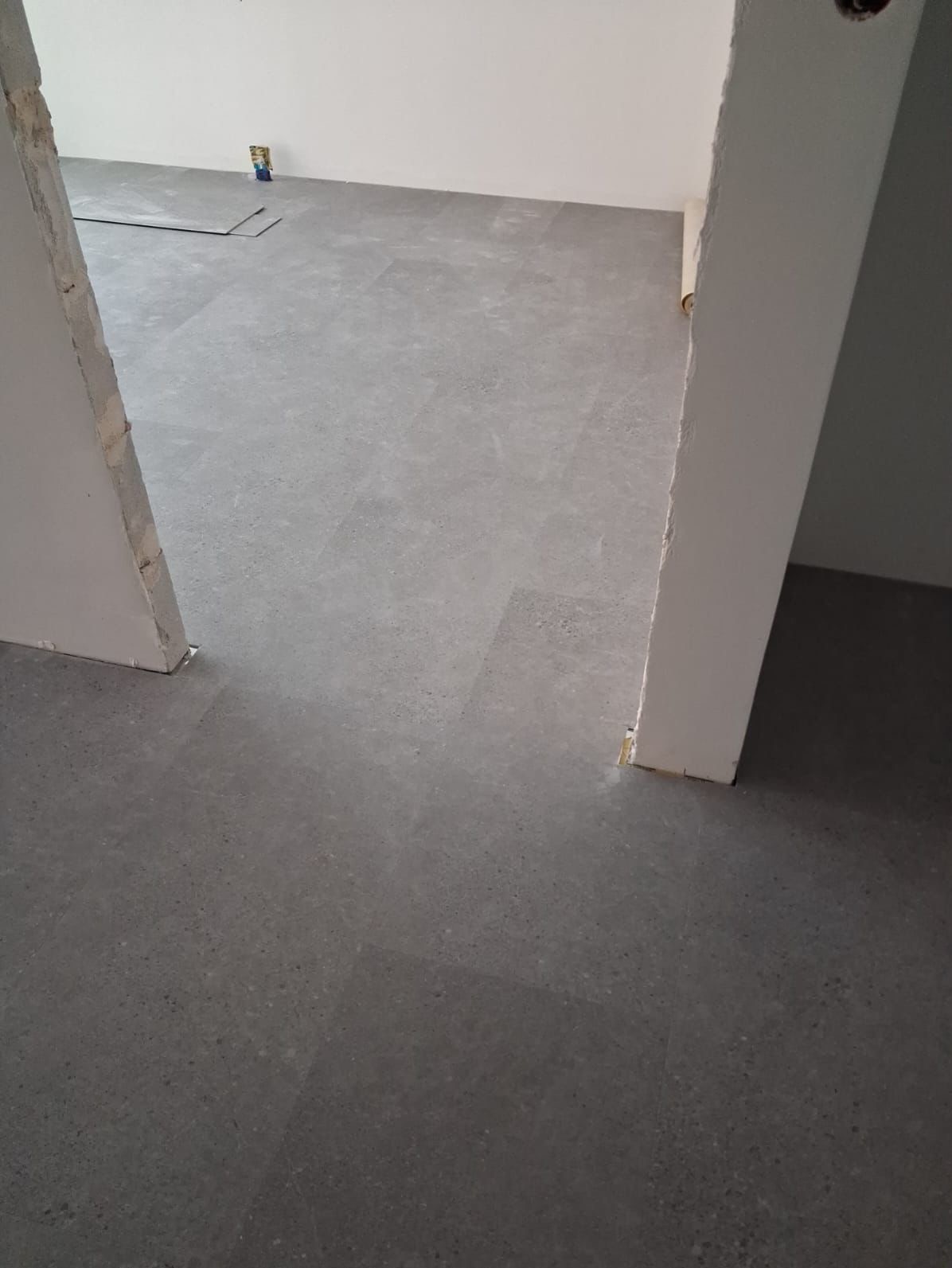 Szara, matowa podłoga z paneli ułożona w nowym pomieszczeniu, widok z perspektywy wejścia, z fragmentem niedokończonego progu i widoczną instalacją elektryczną.