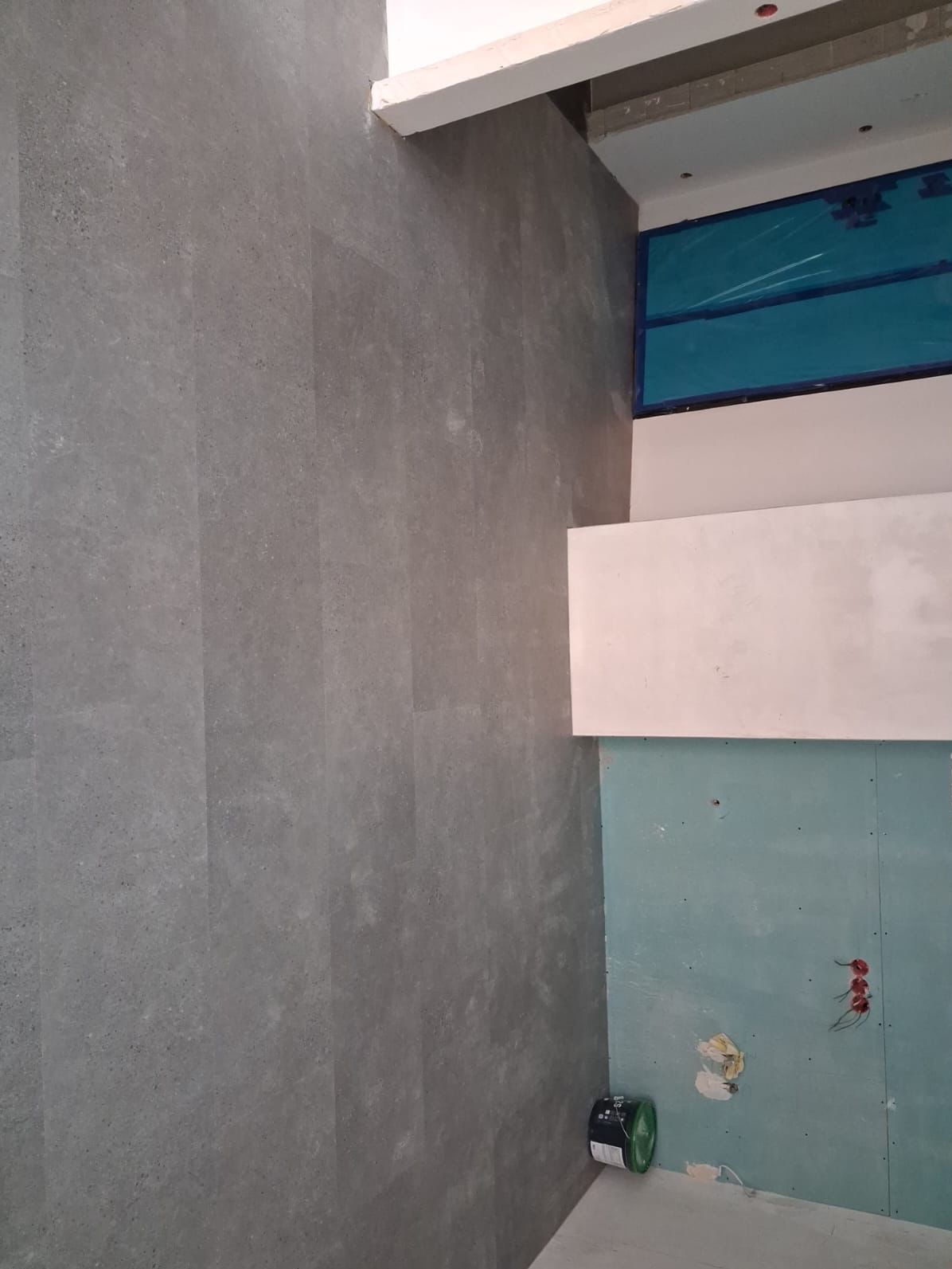 Ściana wyłożona szarymi panelami imitującymi kamień, z widocznymi fugami. Fragment sufitu z niebieską folią ochronną. W rogu puszka farby.