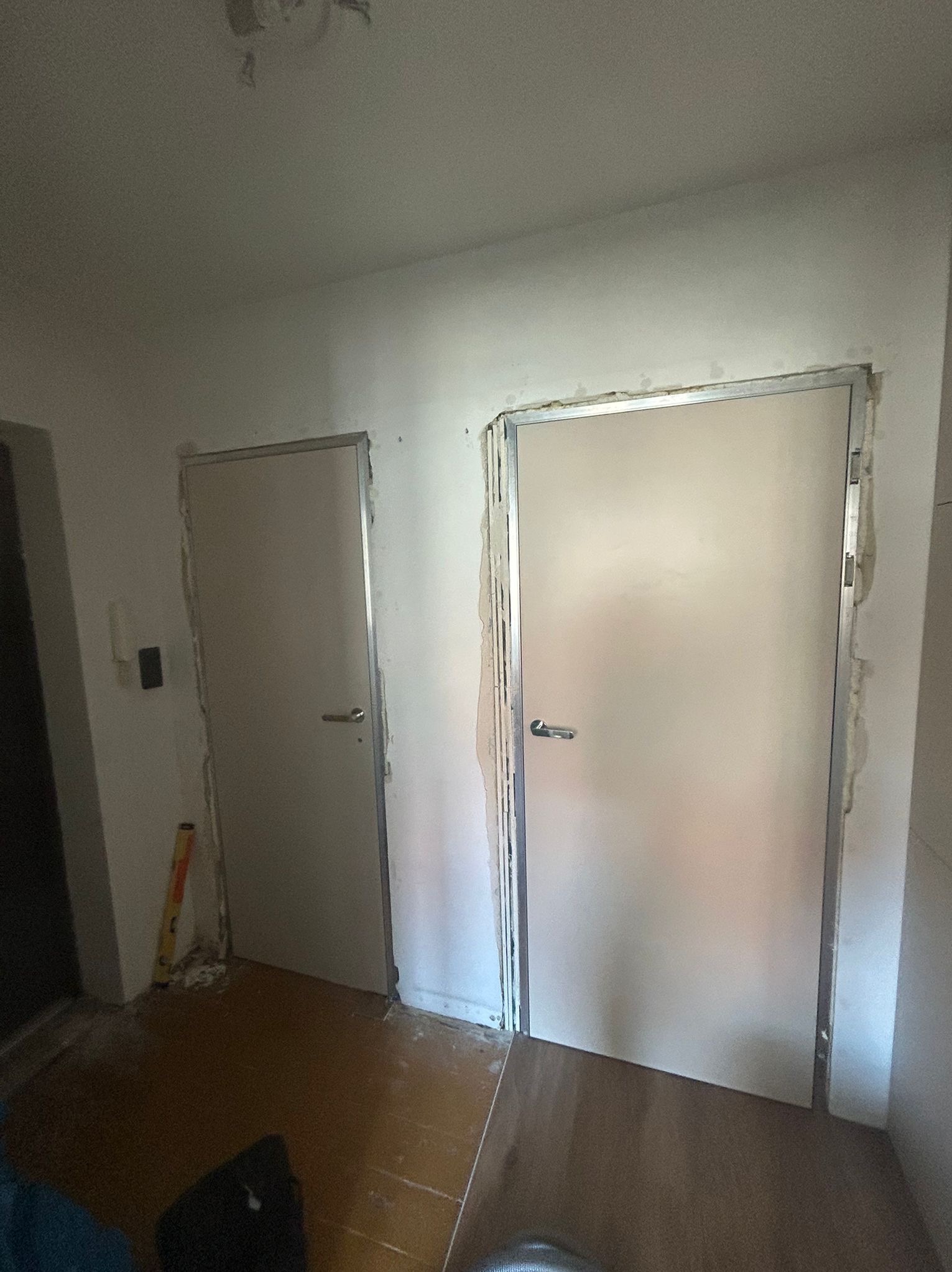 Dwa białe, częściowo zamontowane drzwi z metalowymi ościeżnicami w trakcie prac wykończeniowych. Na podłodze widoczne panele i poziomica.