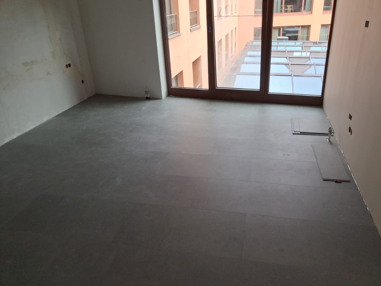 Szare panele podłogowe ułożone w pokoju z dużym oknem. Część paneli jeszcze nieułożona przy ścianie. Widoczne gniazdka elektryczne i fragment budynku za oknem.