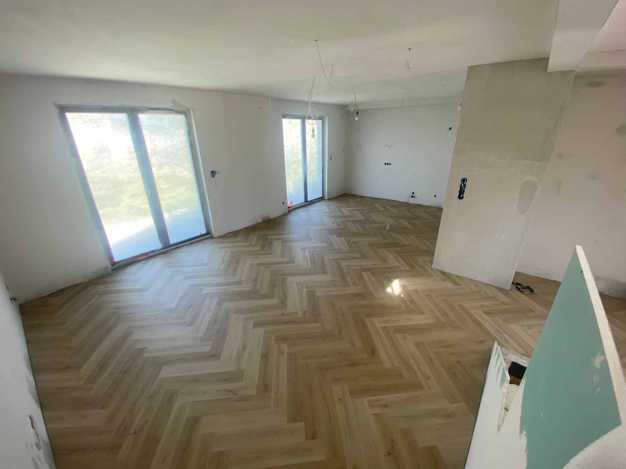 Jasne panele ułożone w jodełkę w przestronnym, jasnym pokoju z dużymi oknami. Ściany w trakcie wykończenia, widoczne kable od oświetlenia.