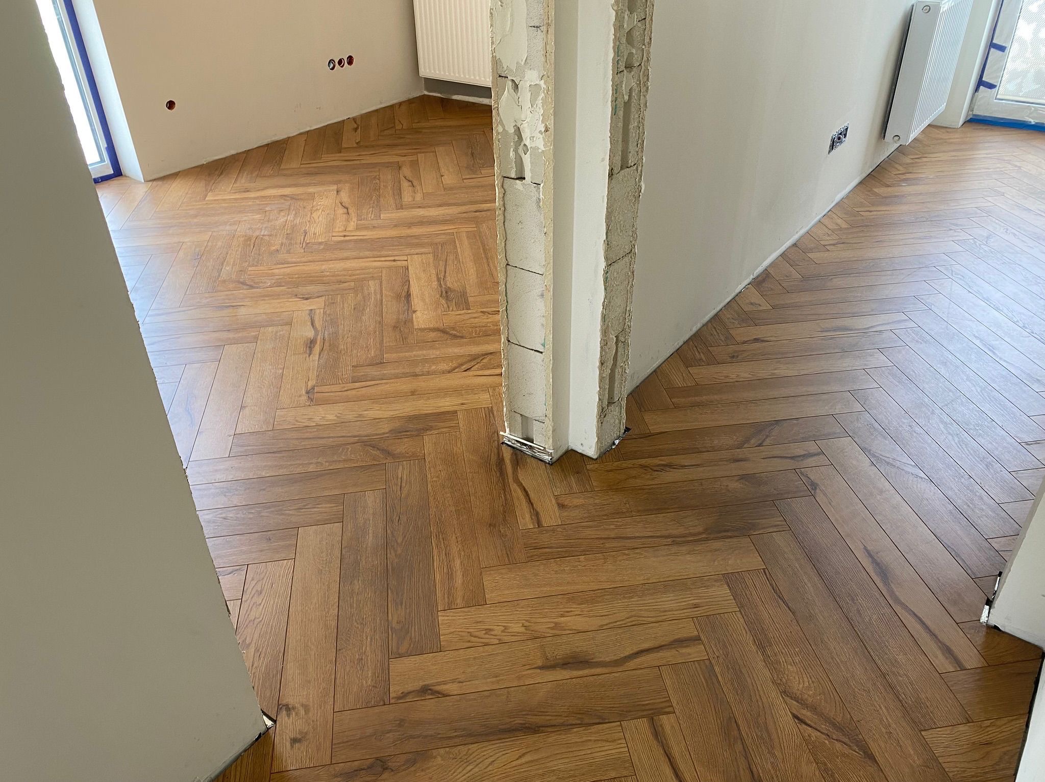 Ukończona podłoga z paneli ułożonych w jodełkę, widoczna w nowym wnętrzu z niedokończonymi ścianami i grzejnikiem. Ciepłe odcienie drewna i wzór jodełki.