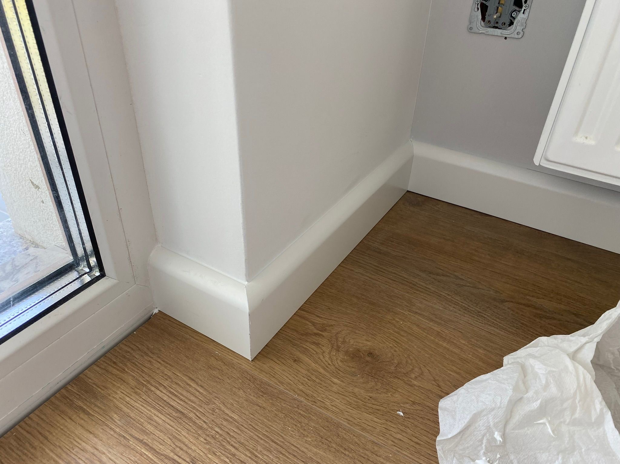 Zamknięty kadr na fragment wnętrza: brązowe panele podłogowe, białe listwy przypodłogowe przy ścianie i oknie. Widoczny fragment gniazdka elektrycznego.