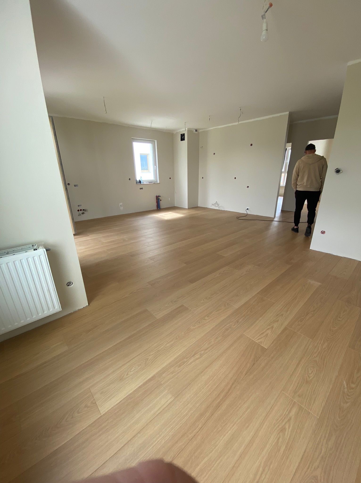 Jasne panele podłogowe w nowym, przestronnym pokoju z grzejnikiem i widocznymi instalacjami elektrycznymi. Osoba w tle przechodzi przez drzwi.