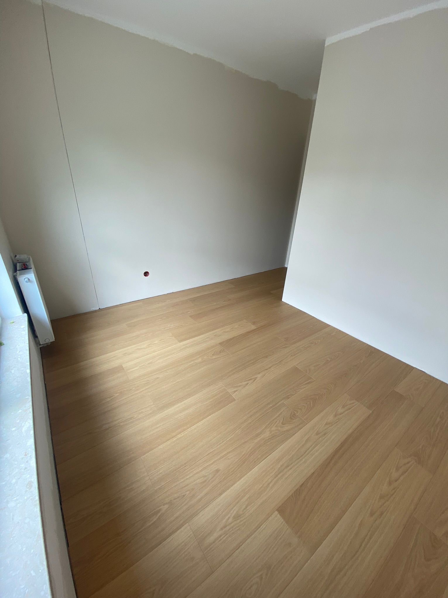 Jasne panele podłogowe ułożone w pustym pokoju z beżowymi ścianami. Widoczny grzejnik przy ścianie i niedokończone wykończenie sufitu.