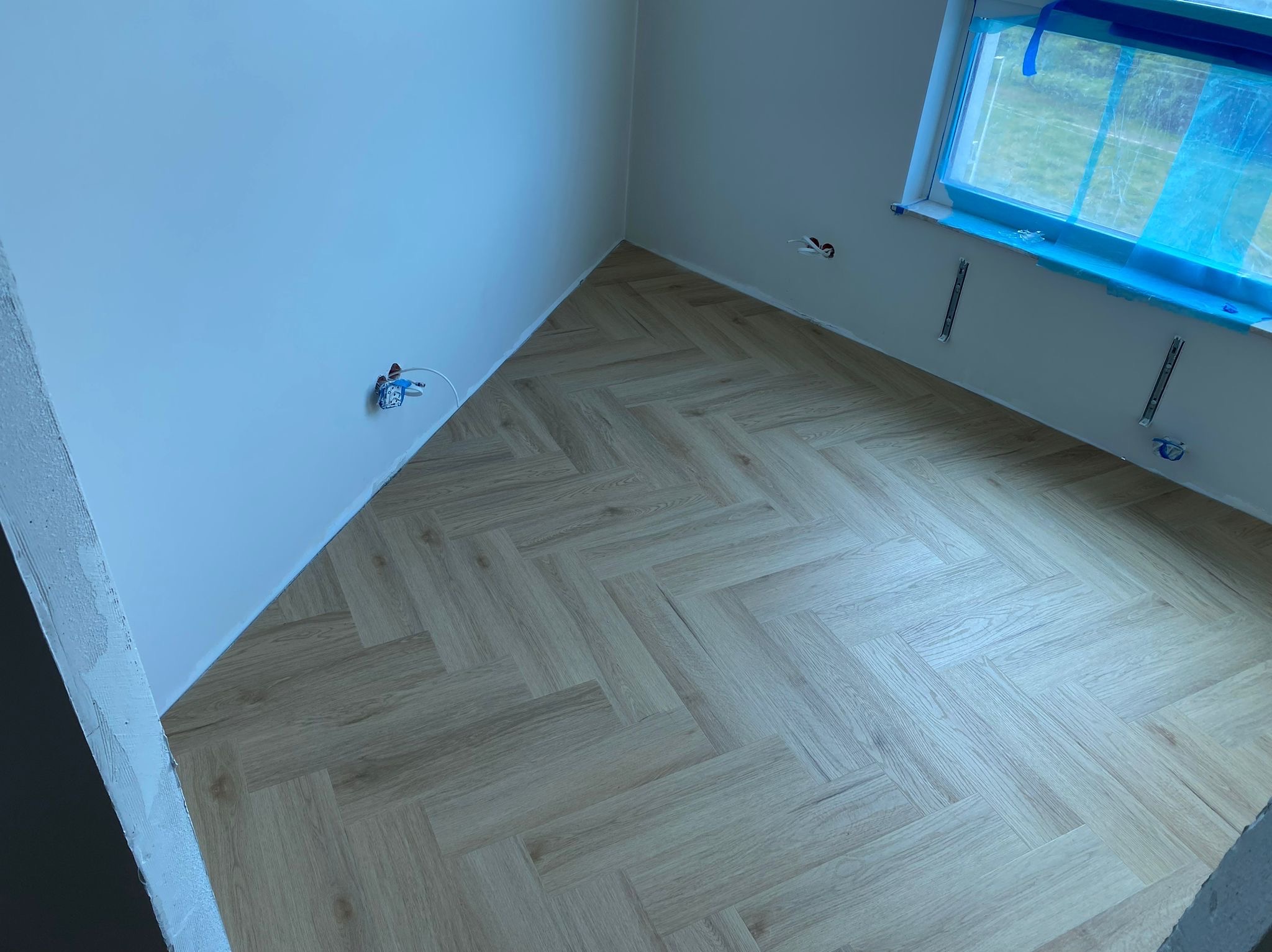 Jasne panele ułożone w jodełkę w rogu pokoju z białymi ścianami i oknem zabezpieczonym folią. Widoczne gniazdka elektryczne i fragment surowego muru.