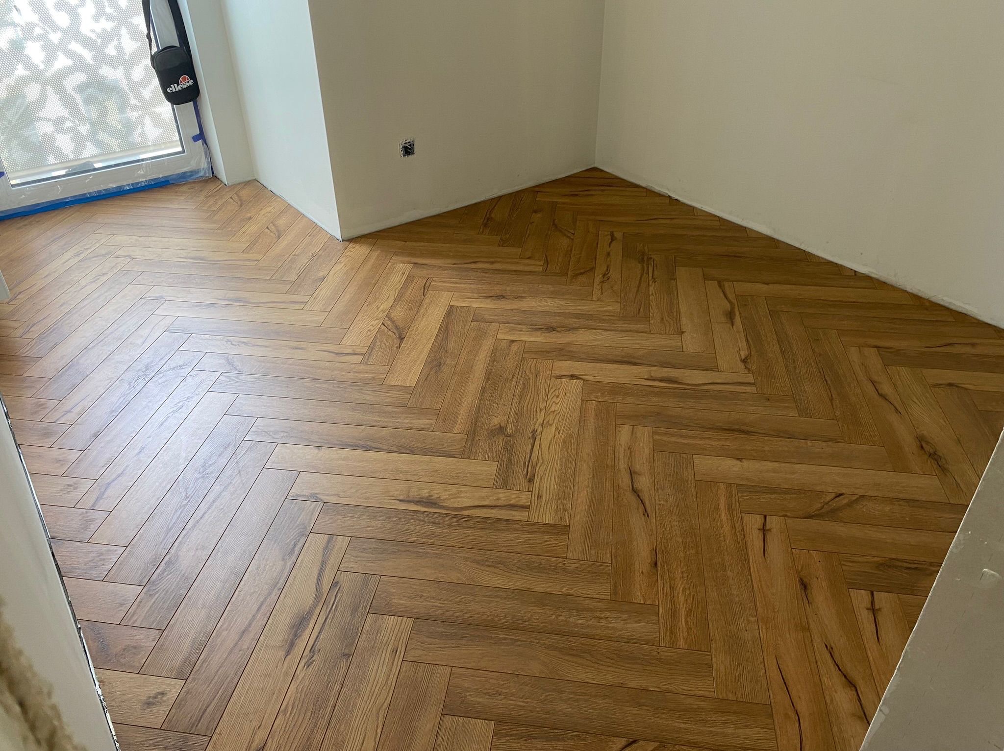 Ułożony parkiet w jodełkę francuską, brązowe panele z widoczną strukturą drewna. Fragment okna z torbą roboczą w rogu. Widok na wykończoną podłogę w pomieszczeniu.