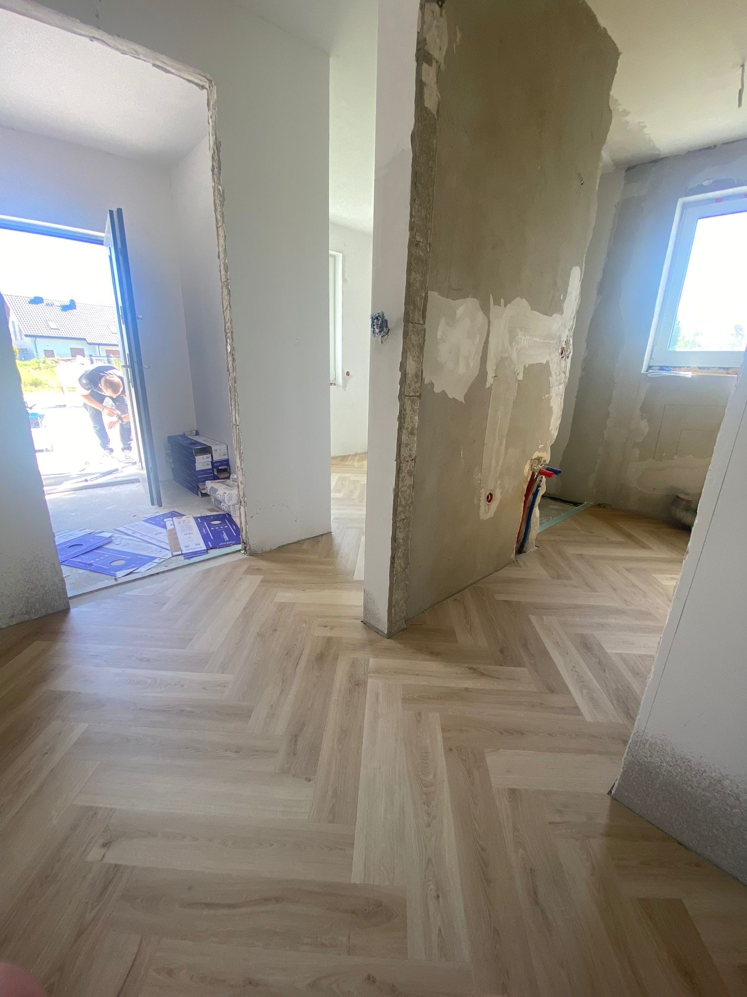 Jasne panele ułożone w jodełkę w surowym wnętrzu. Widoczne ślady po pracach wykończeniowych na ścianach i otwarte przejście do kolejnego pomieszczenia.