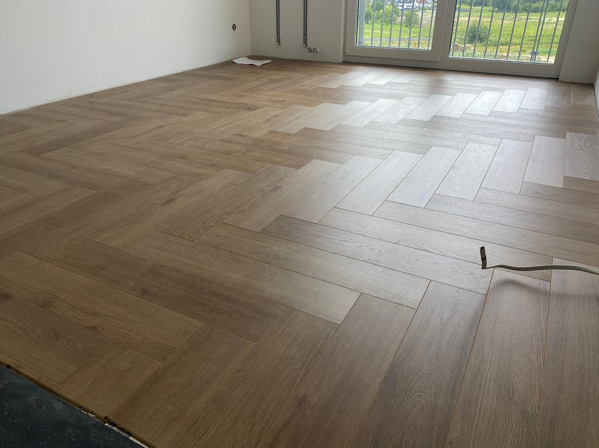 Ułożona podłoga z paneli w jodełkę w jasnym, pustym pokoju z oknem balkonowym. Widoczne fragmenty instalacji grzewczej na ścianie i kabel na podłodze.