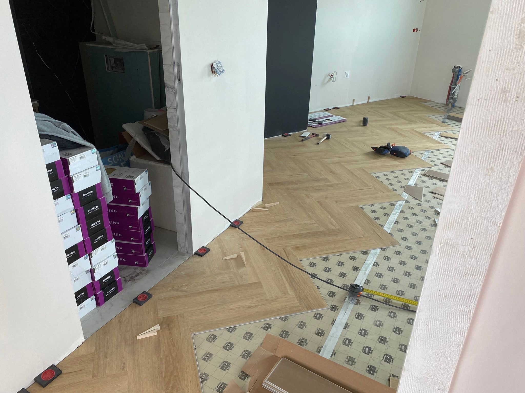 Parkiet w jodełkę układany w Gdańsku. Widoczne narzędzia, listwy przypodłogowe i kartony z panelami. Częściowo ułożona podłoga z jasnego drewna.