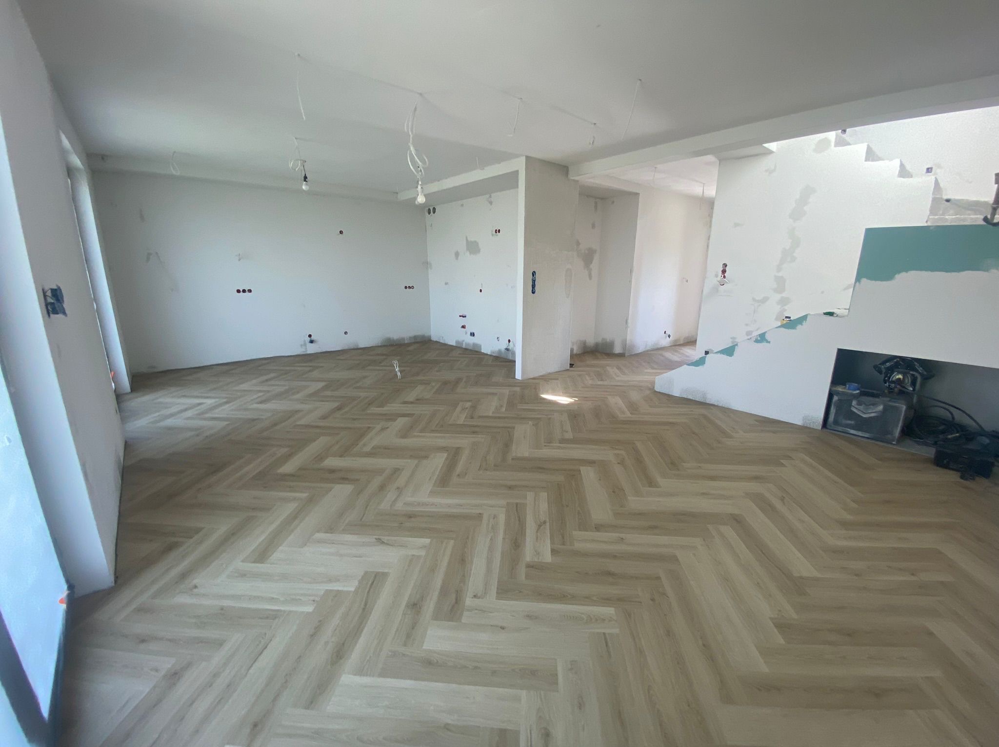 Jasne panele ułożone w jodełkę francuską w przestronnym, surowym wnętrzu. Widoczne niedokończone ściany i instalacje elektryczne. W tle narzędzia.