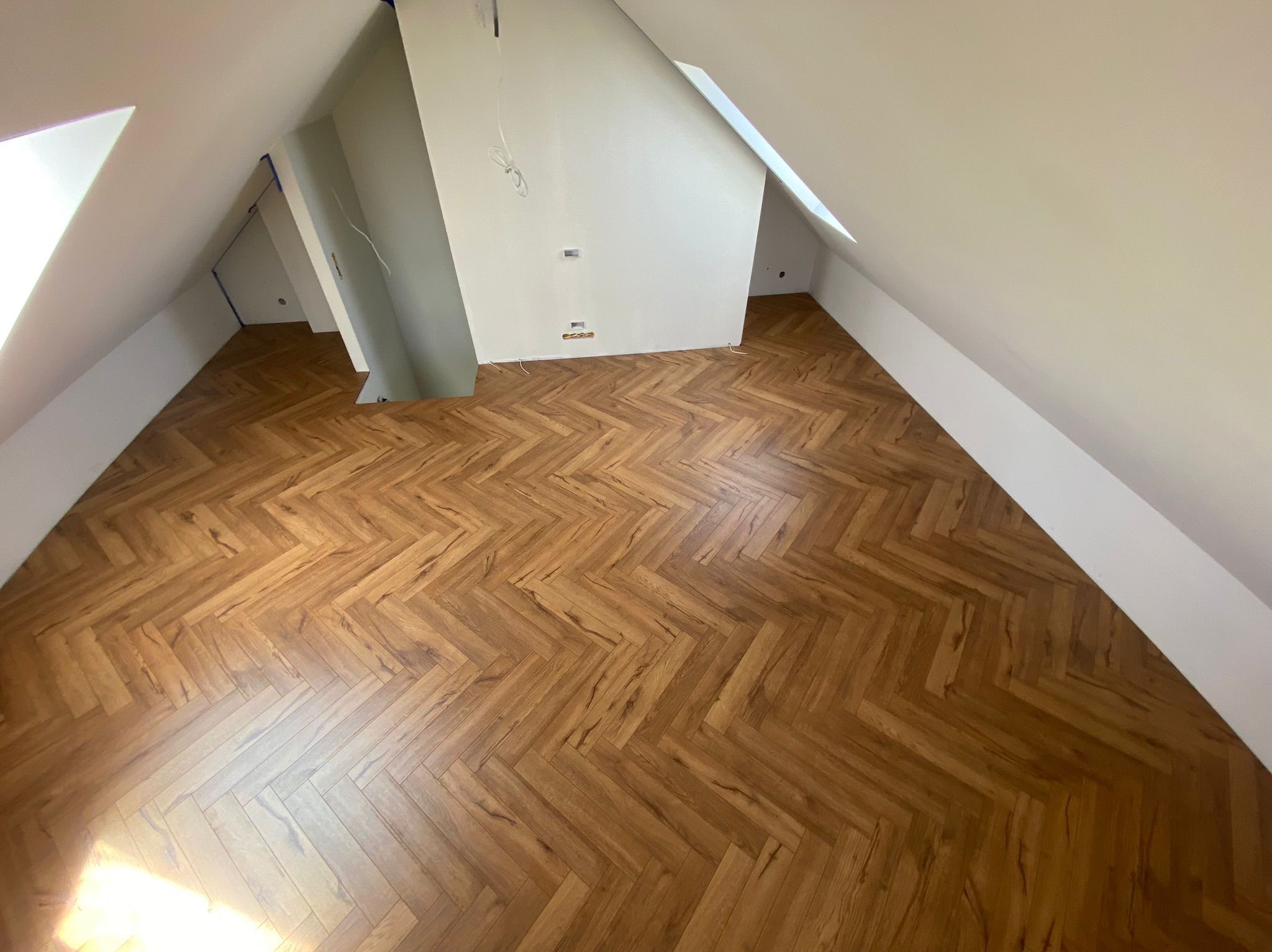 Ukończona podłoga z paneli ułożonych w jodełkę w jasnym, przestronnym pokoju na poddaszu. Widoczne białe ściany i skosy dachu. Kabel wystaje ze ściany.