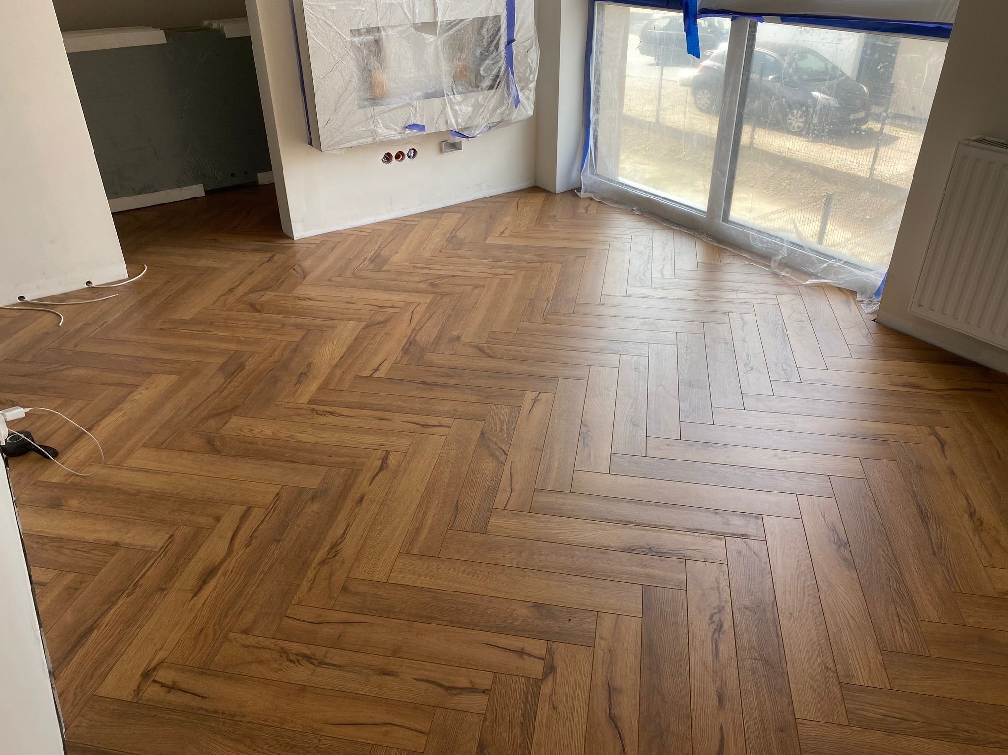 Ukończona podłoga z paneli ułożonych w jodełkę, z widocznym zabezpieczeniem okna i ściany folią malarską. Ciepłe odcienie drewna i naturalne światło.