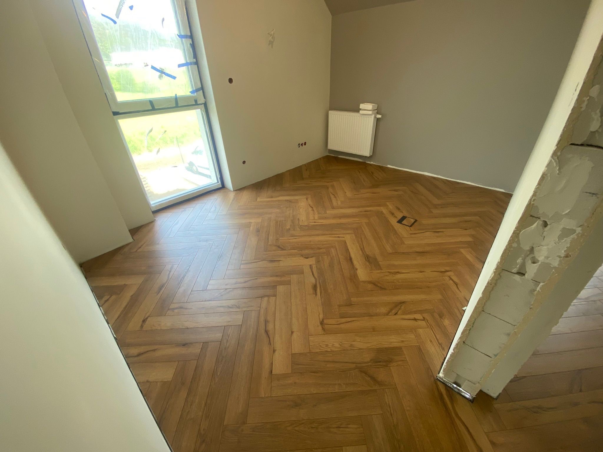 Ukończona podłoga z paneli ułożonych w jodełkę w jasnym pokoju z oknem i grzejnikiem. Widoczny fragment futryny drzwi.