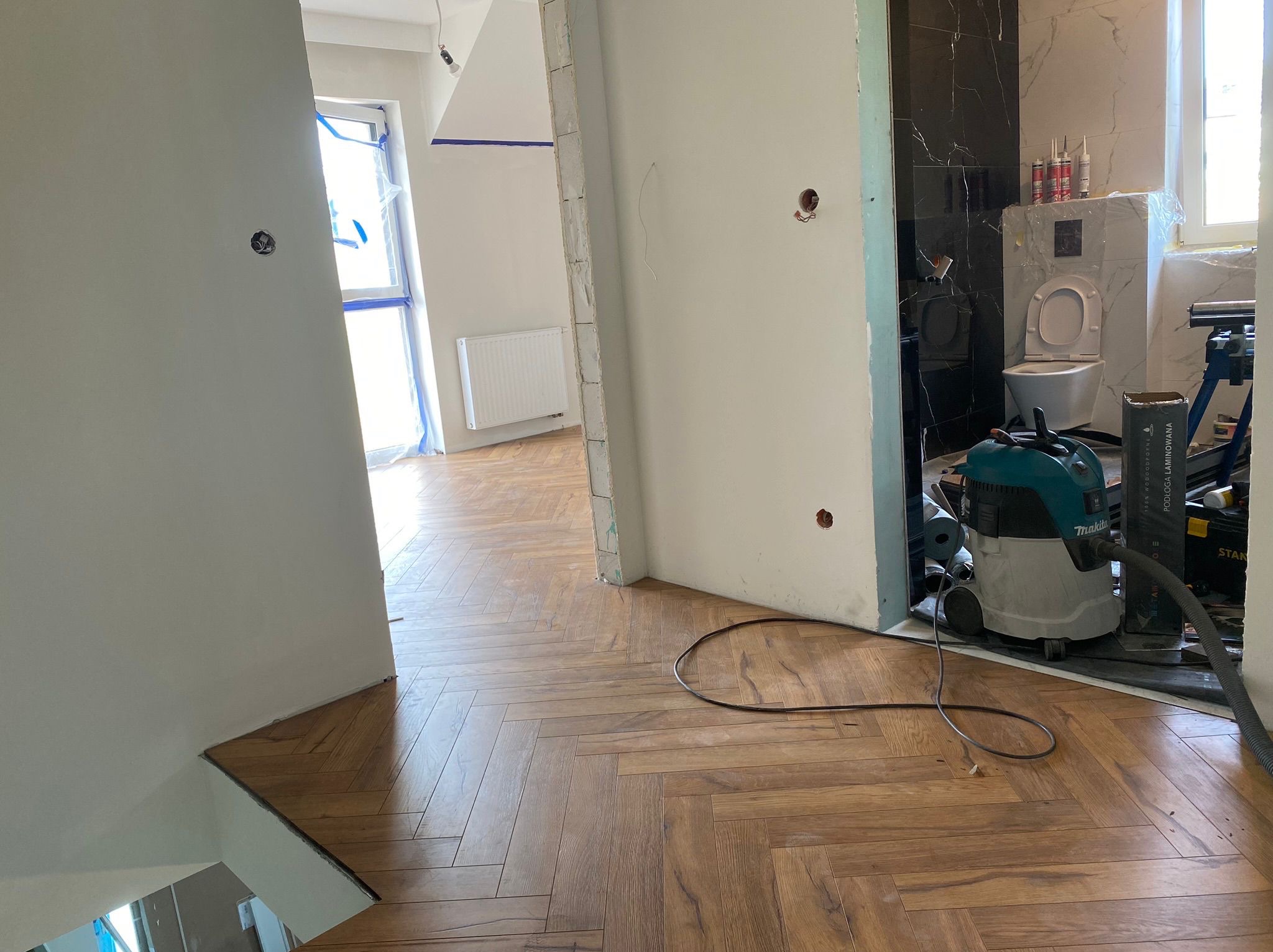 Ukończona podłoga z paneli ułożonych w jodełkę, widoczny odkurzacz budowlany i fragment łazienki w trakcie wykańczania. Jasne wnętrze w remoncie.