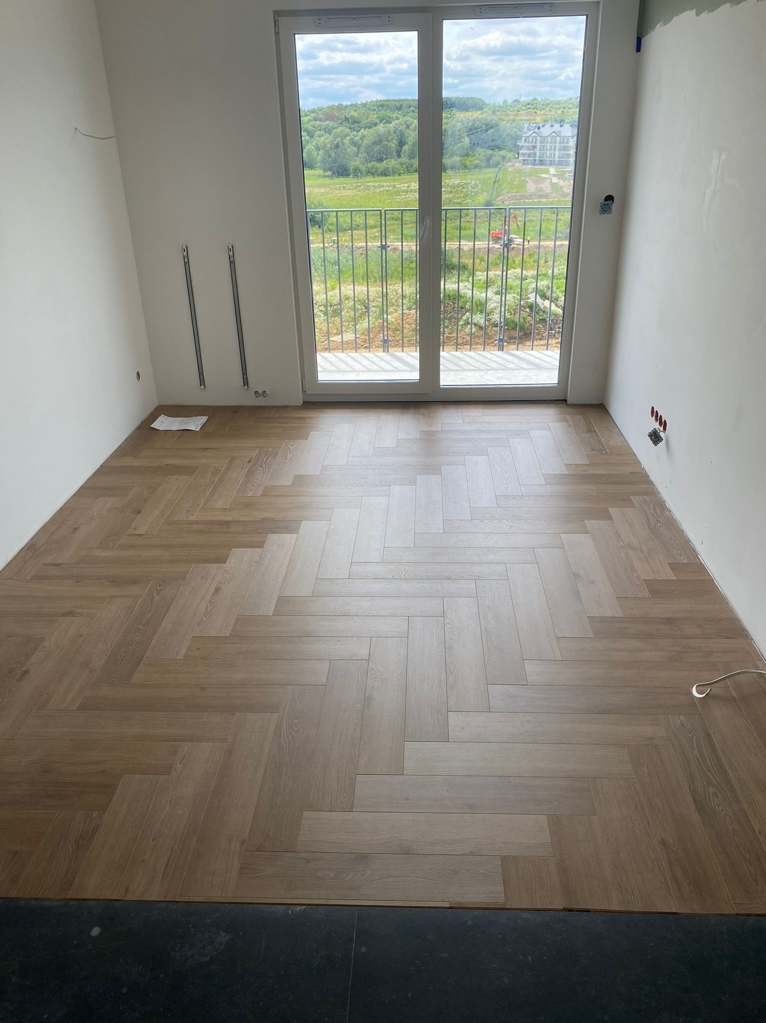 Jasne panele ułożone w jodełkę francuską w pokoju z balkonem. Widoczne niedokończone ściany i wystające kable. Za oknem zielony krajobraz i budynki.