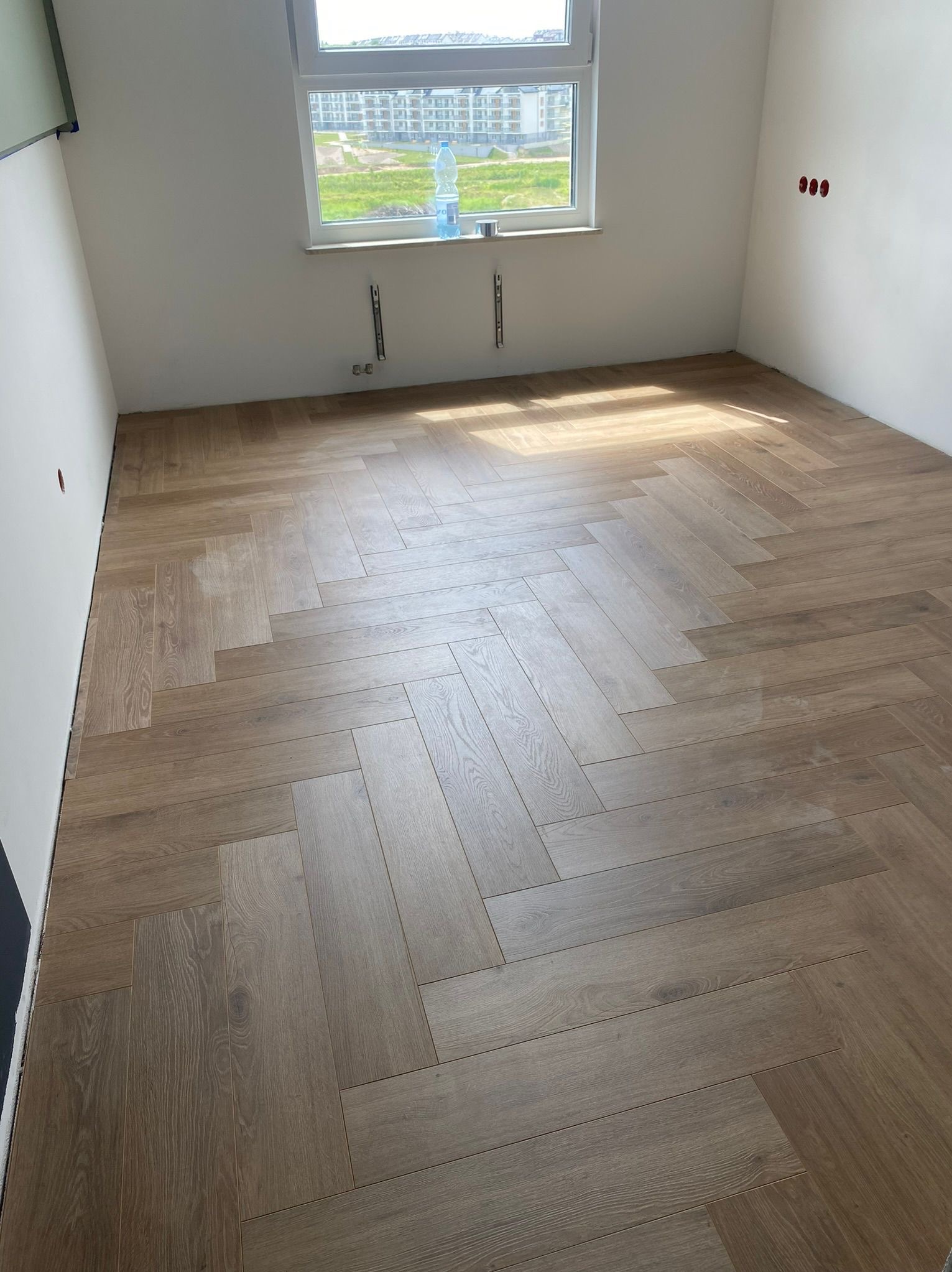 Jasne panele ułożone w jodełkę w pustym pokoju z oknem. Widoczne niedokończone ściany i wystające kable. Naturalne światło podkreśla wzór podłogi.