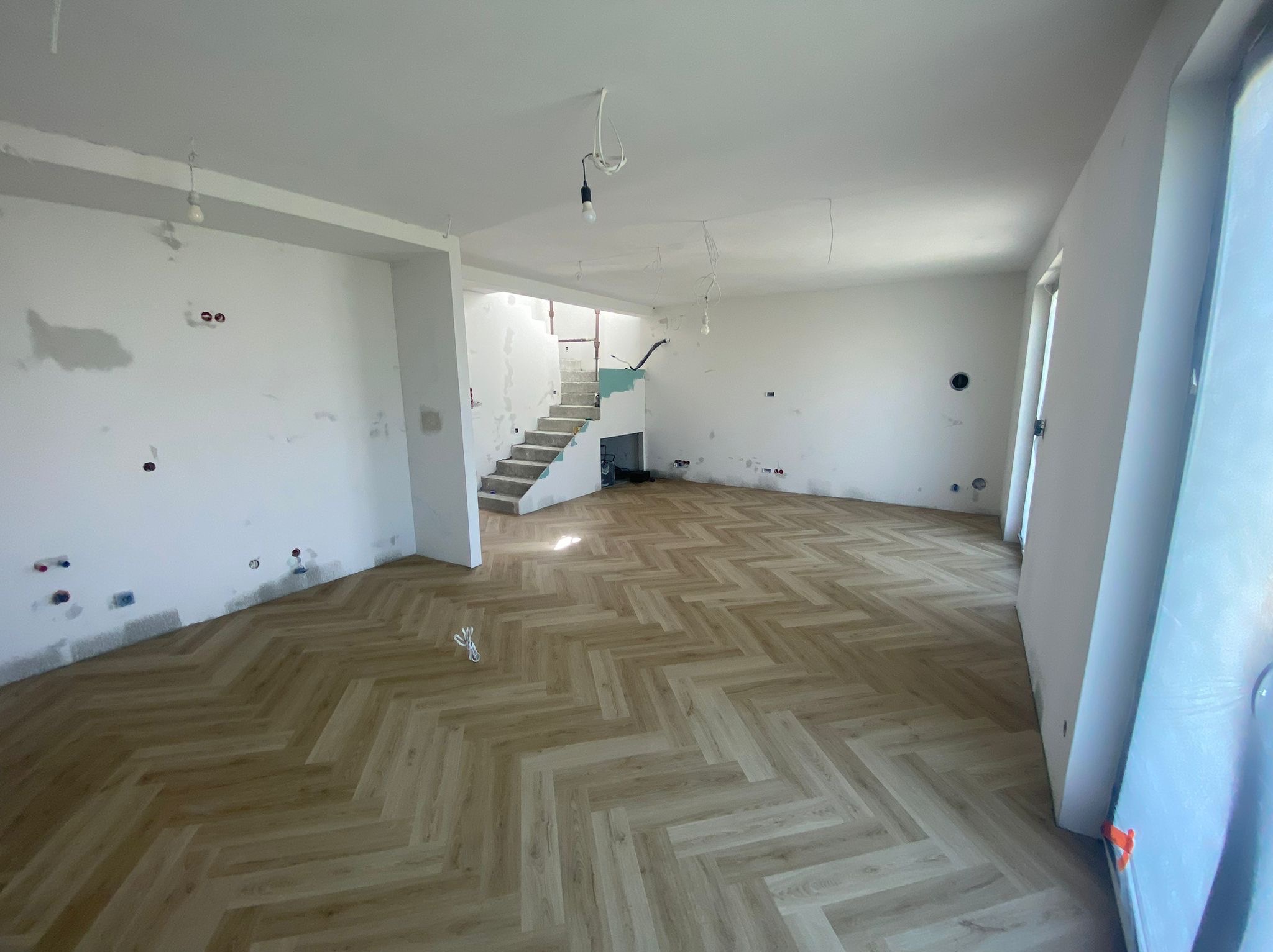 Jasne panele ułożone we wzór jodełki w przestronnym, ale jeszcze surowym wnętrzu. Widoczne niedokończone ściany i schody. Nowa podłoga kontrastuje z resztą pomieszczenia.