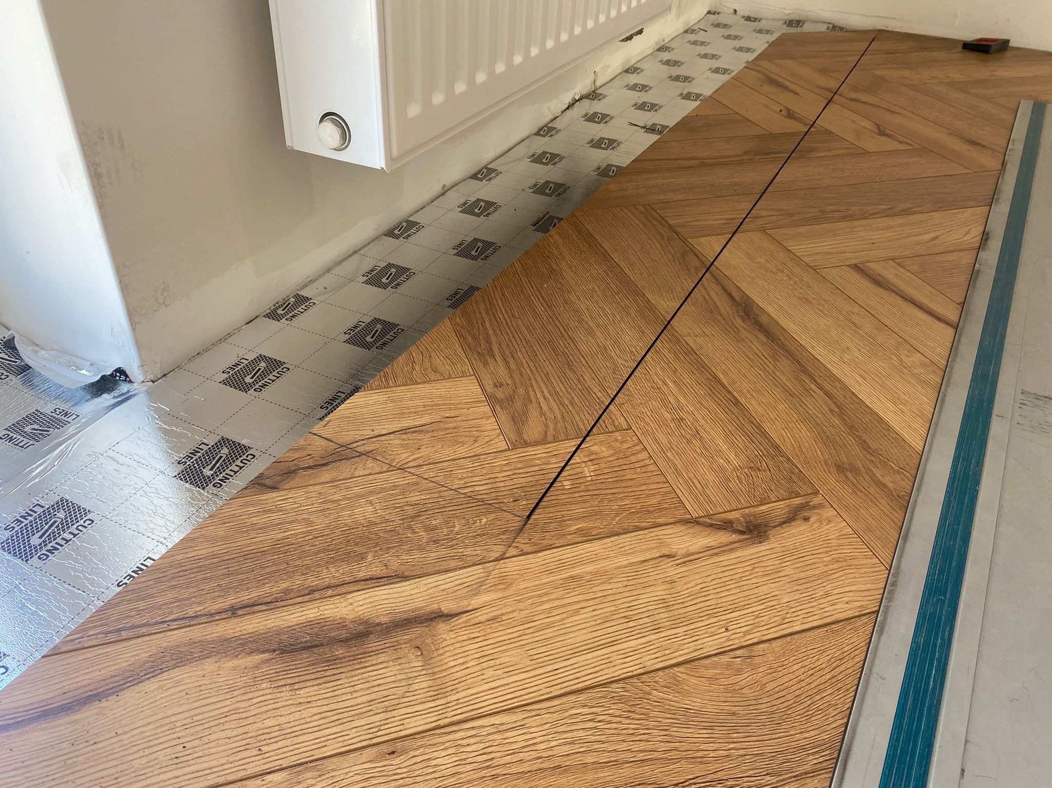 Układanie parkietu w jodełkę francuską, widoczna folia izolacyjna, grzejnik i listwa przypodłogowa. Częściowo ułożona podłoga z drewnianych paneli.