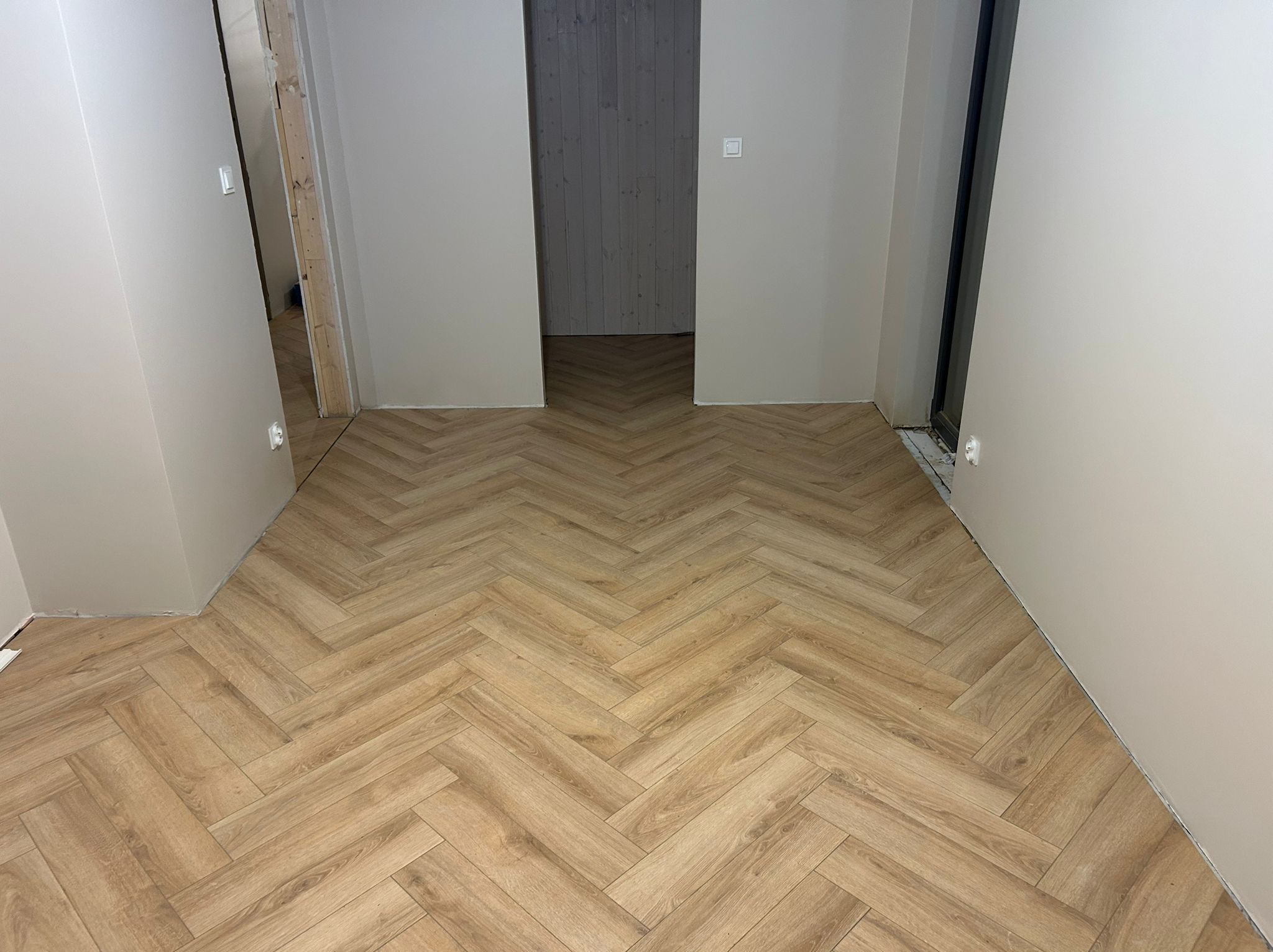 Jasne panele ułożone w jodełkę w korytarzu z widocznymi ościeżnicami drzwiowymi. Ściany w odcieniach beżu. Widok na sąsiednie pomieszczenie z szarą ścianą.