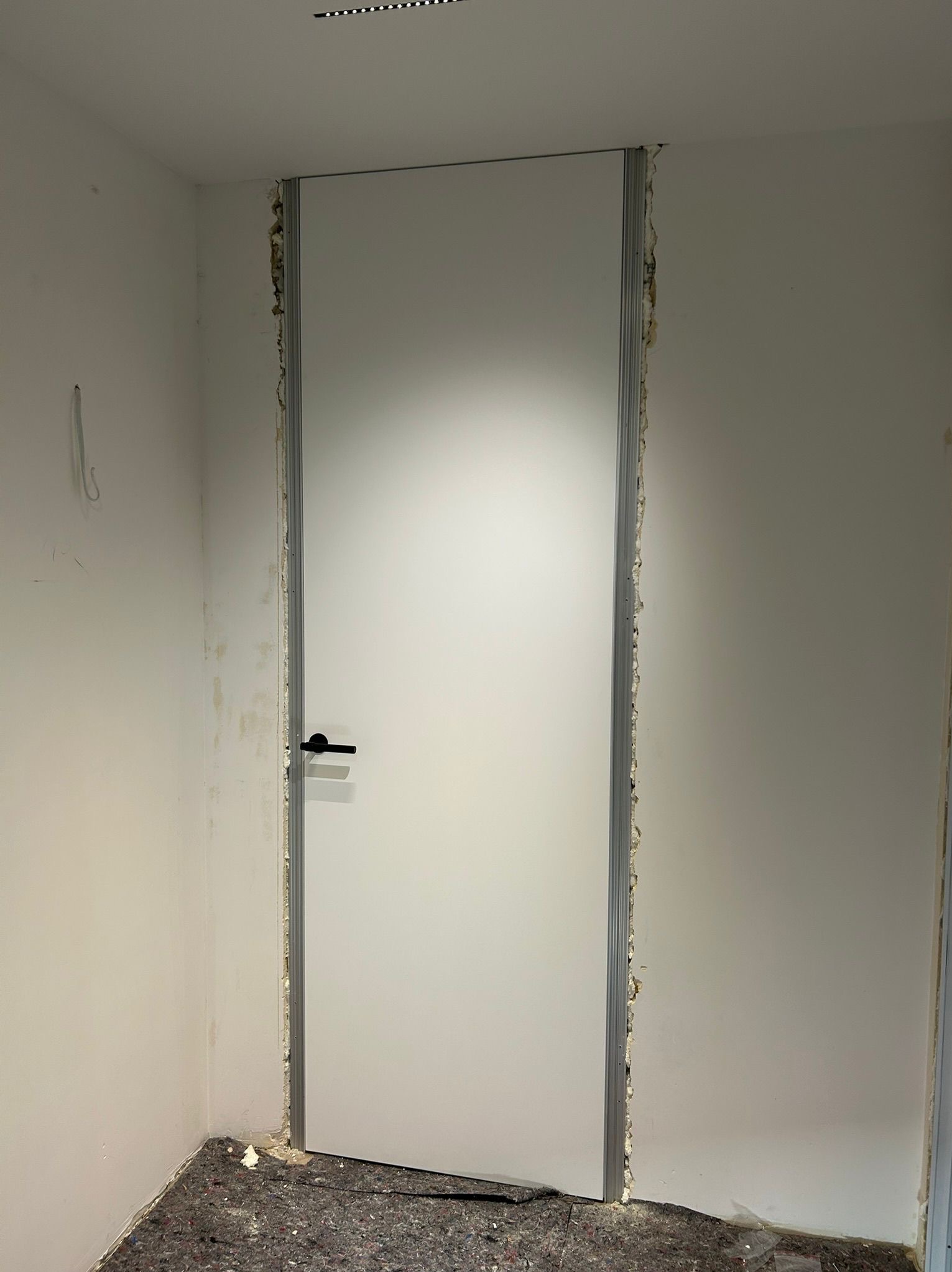 Białe drzwi z czarną klamką, osadzone w surowym otworze, widoczne pianka montażowa i niedokończone wykończenie ścian. Podłoga zabezpieczona materiałem.