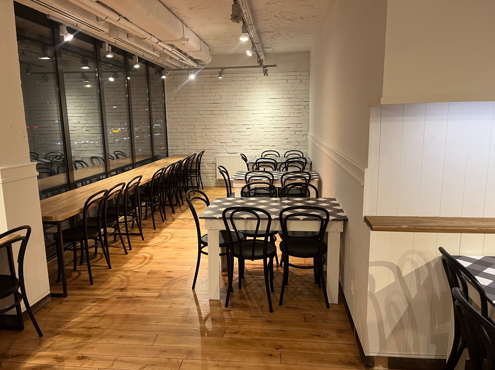 Jasne wnętrze biura z długim stołem wzdłuż okna i stolikami z krzesłami. Parkiet na podłodze, biała cegła na ścianie. Oświetlenie sufitowe.