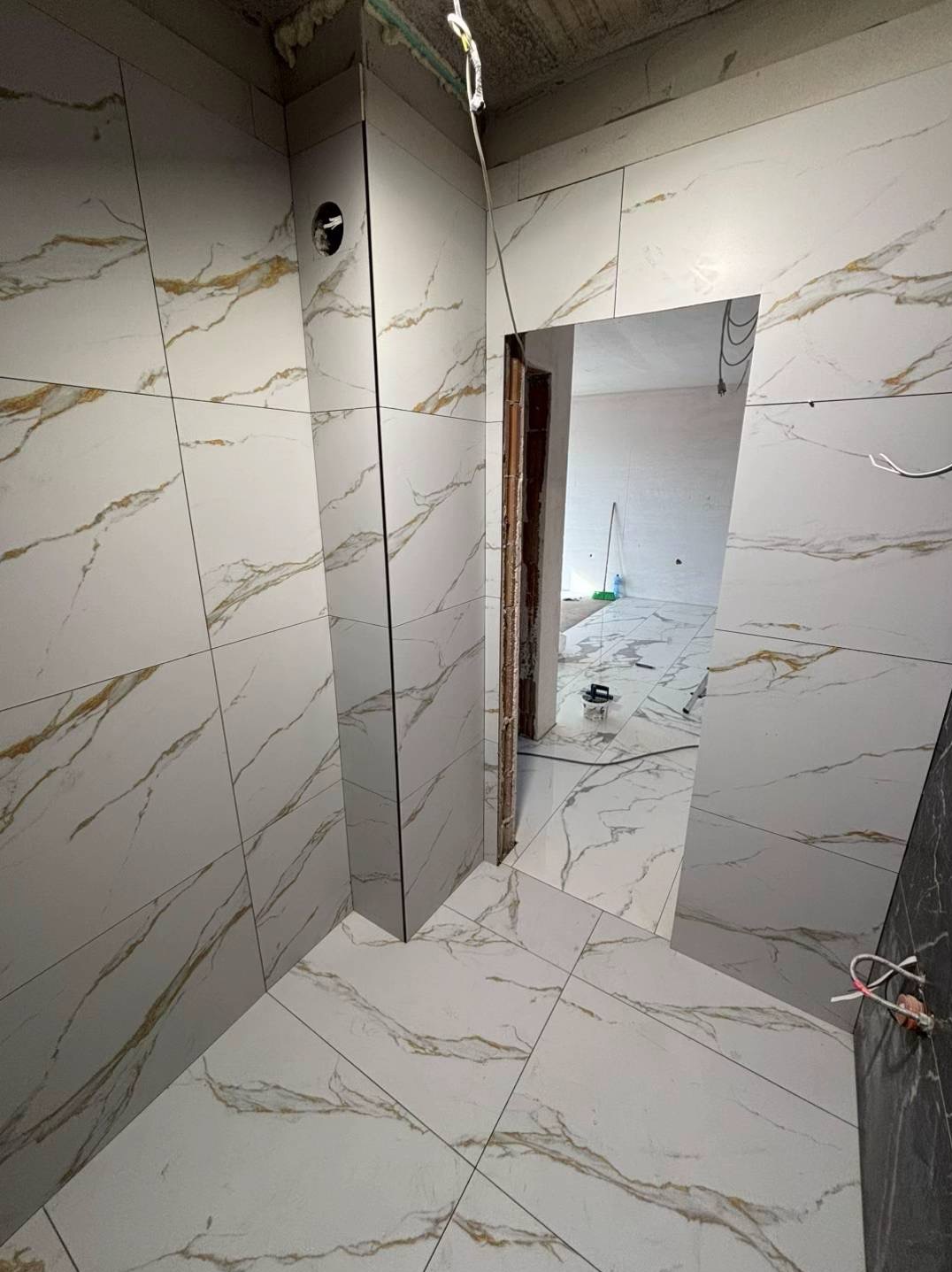 Białe płytki imitujące marmur z złotymi żyłami, ułożone na ścianach i podłodze łazienki w trakcie remontu. Widoczne narzędzia i niedokończone wykończenia.