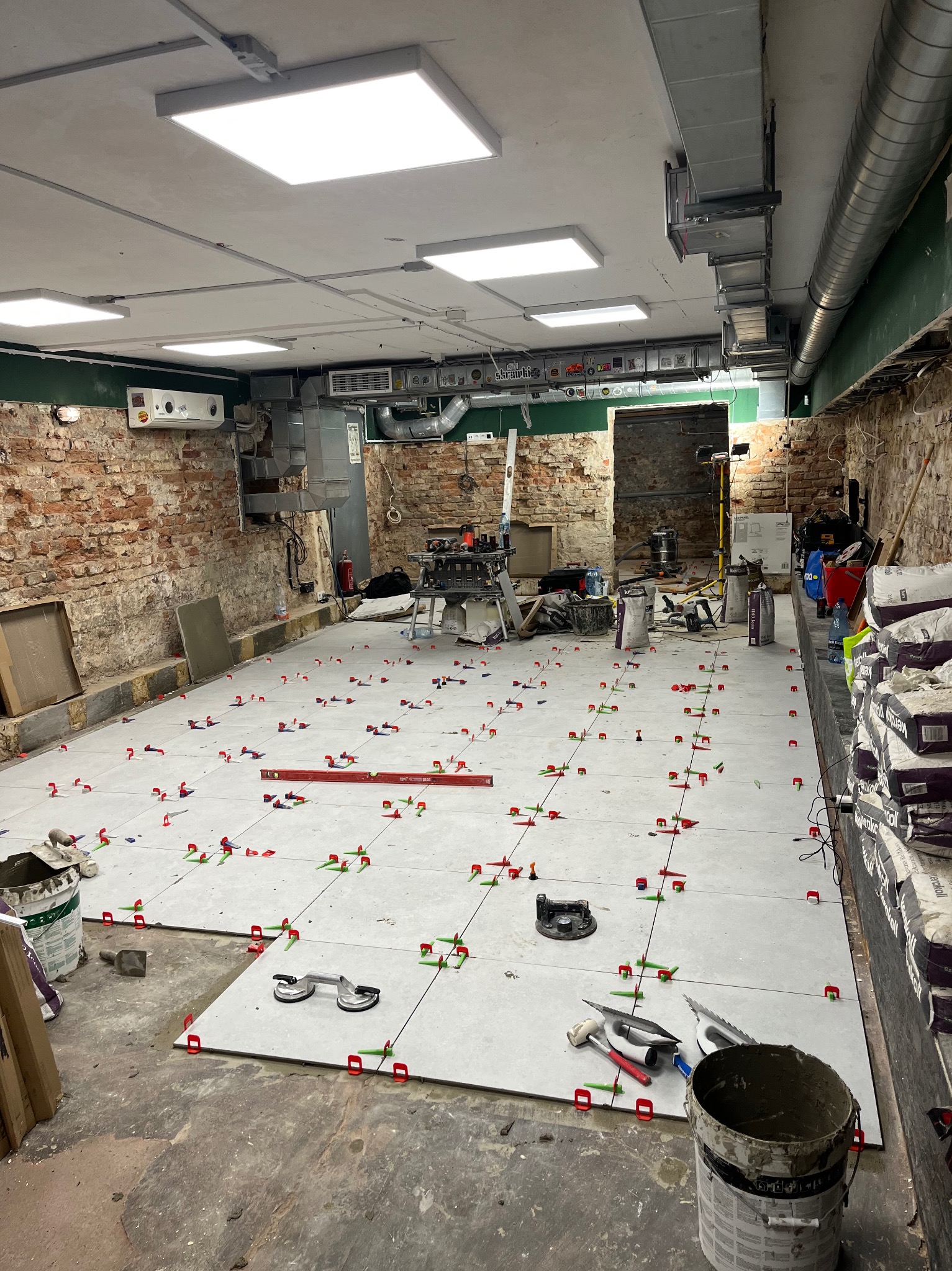 Układanie jasnych płytek podłogowych z użyciem systemu poziomowania. Widoczne narzędzia glazurnicze i worki z zaprawą. Ceglana ściana w tle, surowy stan.