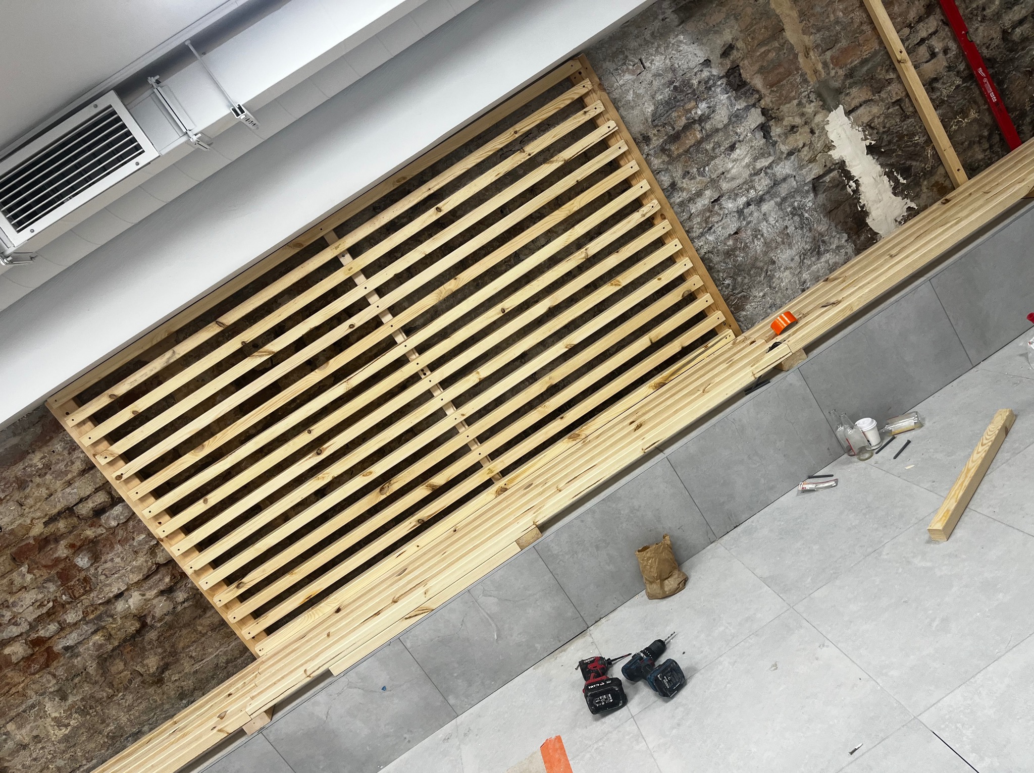 Montaż drewnianej konstrukcji na ścianie z cegły w łazience. Widoczne narzędzia i płytki podłogowe. Prace wykończeniowe w trakcie realizacji.