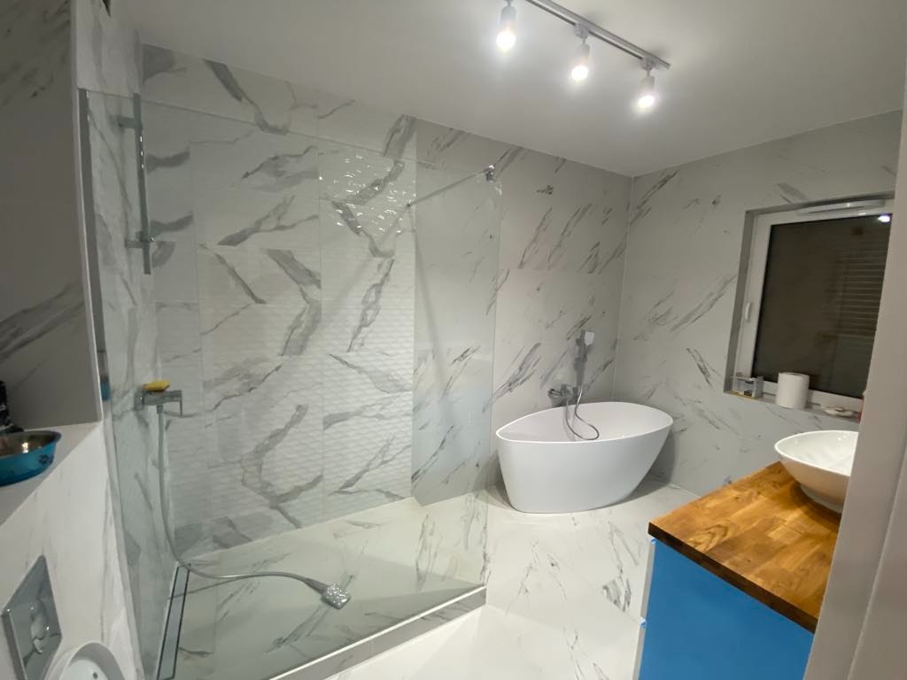 Nowoczesna łazienka z kabiną prysznicową typu walk-in, wolnostojącą wanną i umywalką na drewnianym blacie. Białe płytki imitujące marmur na ścianach i podłodze.