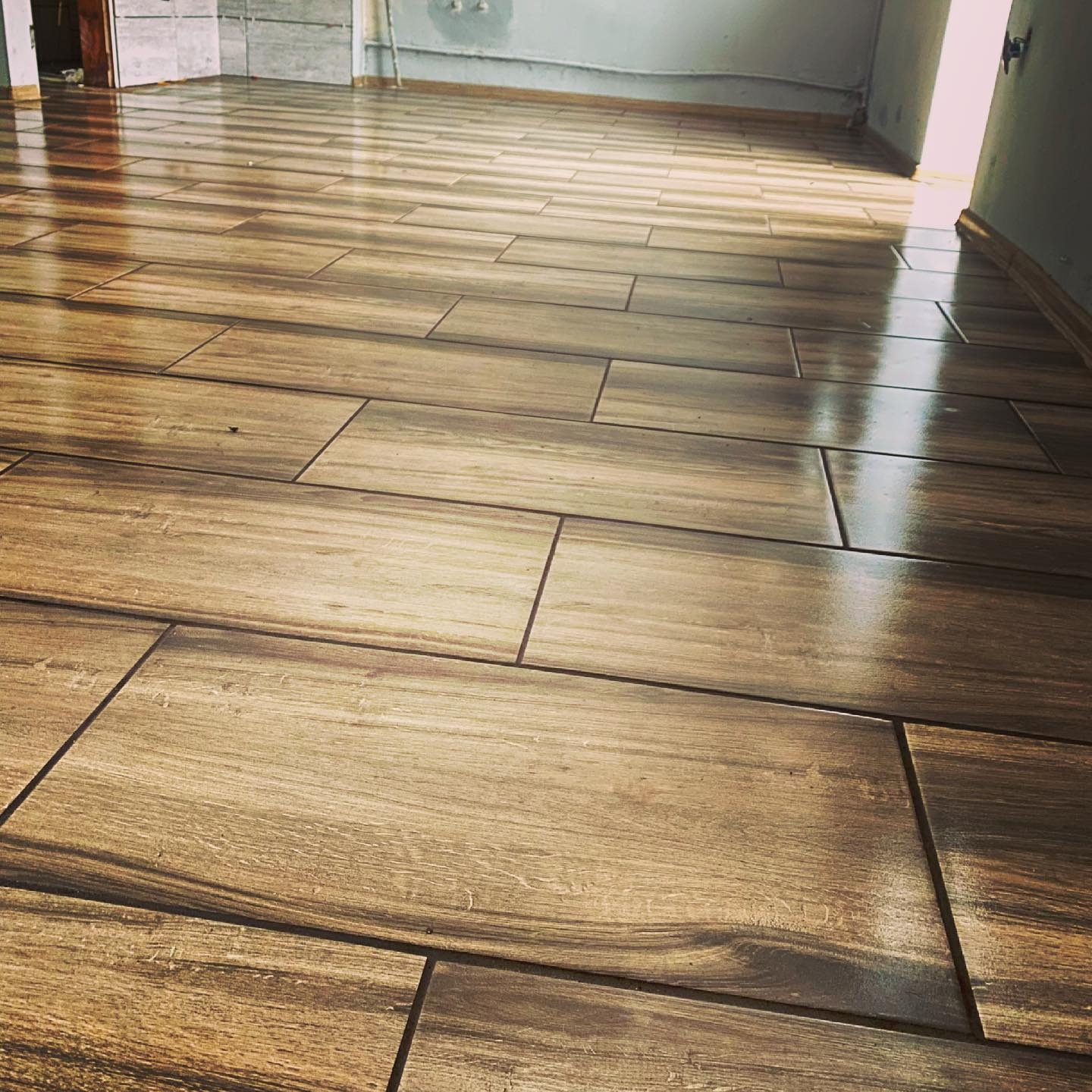 Połyskująca, drewnopodobna podłoga z płytek, ułożona w prostokątnym wzorze. Widoczne fugi i fragmenty ścian w surowym stanie. Perspektywa od wejścia do pokoju.