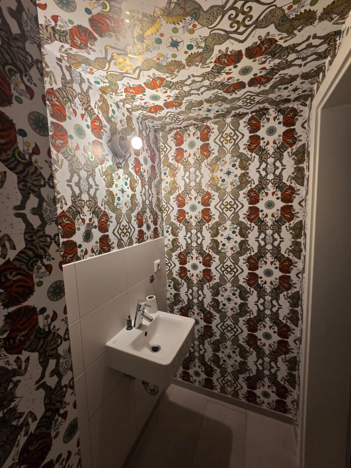 Nietypowa łazienka z wzorzystą tapetą w motywy zwierzęce na ścianach i suficie. Biała umywalka na tle płytek. Oświetlenie punktowe.