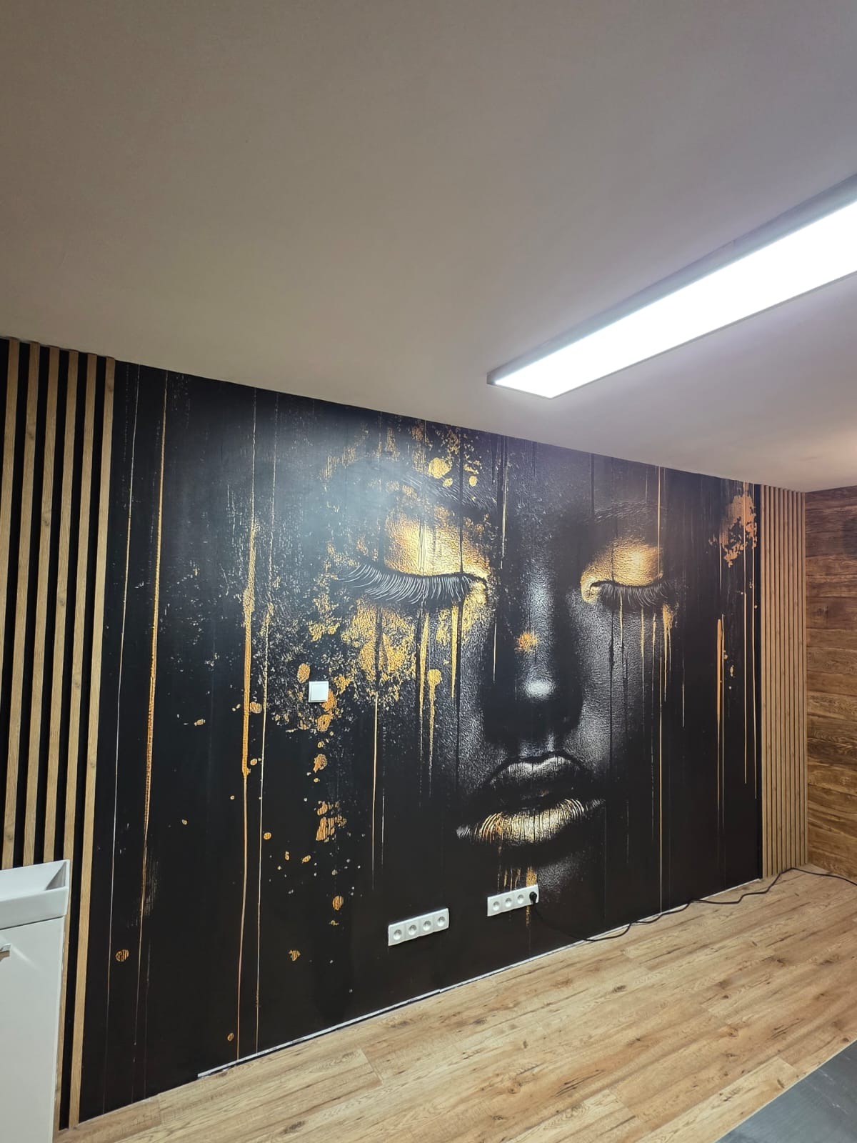 Nowoczesne wykończenie wnętrza: ściana z fototapetą z motywem twarzy w czerni i złocie, panele pionowe z drewna, jasna podłoga i oświetlenie sufitowe.