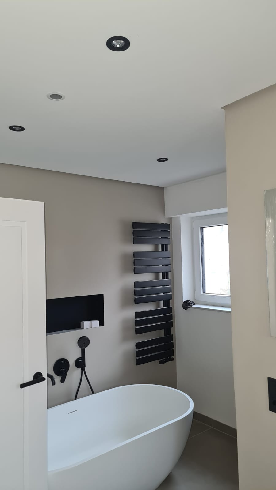 Minimalistyczna łazienka z wolnostojącą wanną, czarną armaturą i grzejnikiem drabinkowym. Oświetlenie punktowe w suficie. Neutralna kolorystyka ścian.