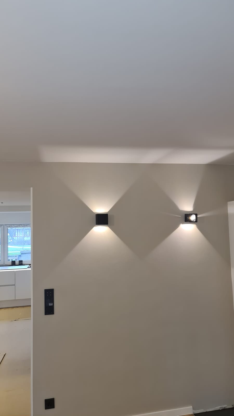 Dwa czarne kinkiety ścienne oświetlają beżową ścianę w nowocześnie wykończonym wnętrzu. Widoczne włączniki i fragment kuchni w tle.