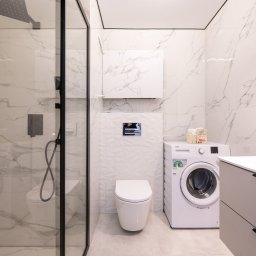 Usługi budowlane - Nowoczesna łazienka z kabiną prysznicową typu walk-in, toaletą, pralką i szafką pod umywalką. Białe marmurowe płytki na ścianach i szare meble.