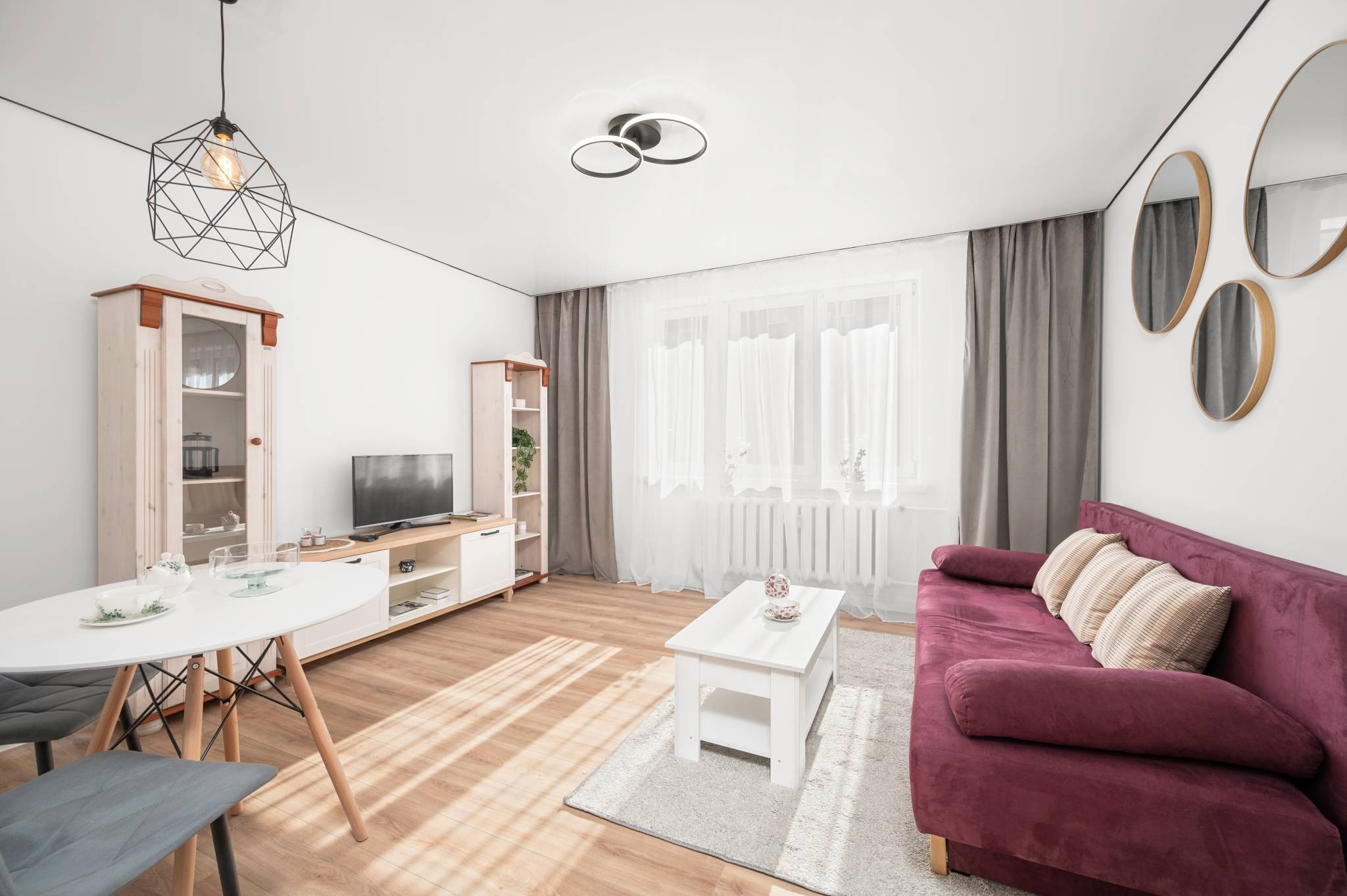 Jasne wnętrze salonu z drewnianą podłogą, burgundową sofą i białymi meblami. Okrągłe lustra na ścianie i designerskie lampy dodają nowoczesnego charakteru.