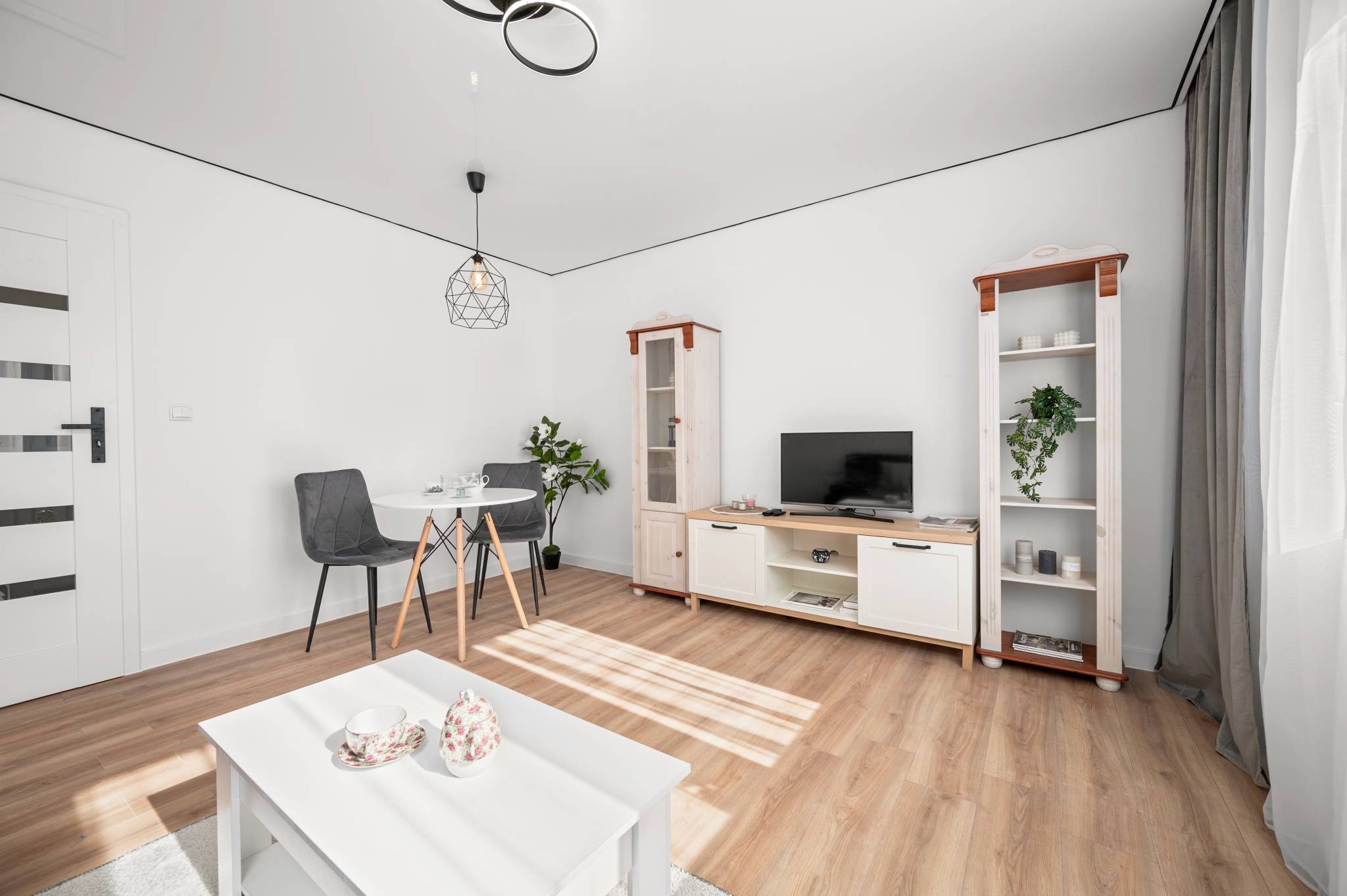 Jasne wnętrze po remoncie: białe ściany z czarnym akcentem, drewniana podłoga, nowoczesne meble i oświetlenie. Minimalistyczny design z roślinami doniczkowymi.