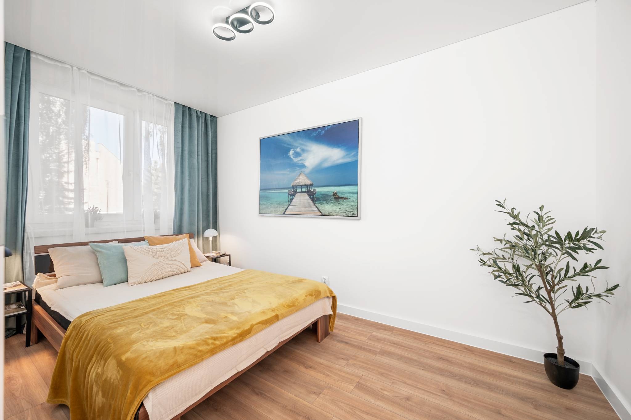 Jasna sypialnia z drewnianą podłogą, łóżkiem z żółtą narzutą, obrazem z motywem morskim i rośliną doniczkową. Minimalistyczne oświetlenie sufitowe.